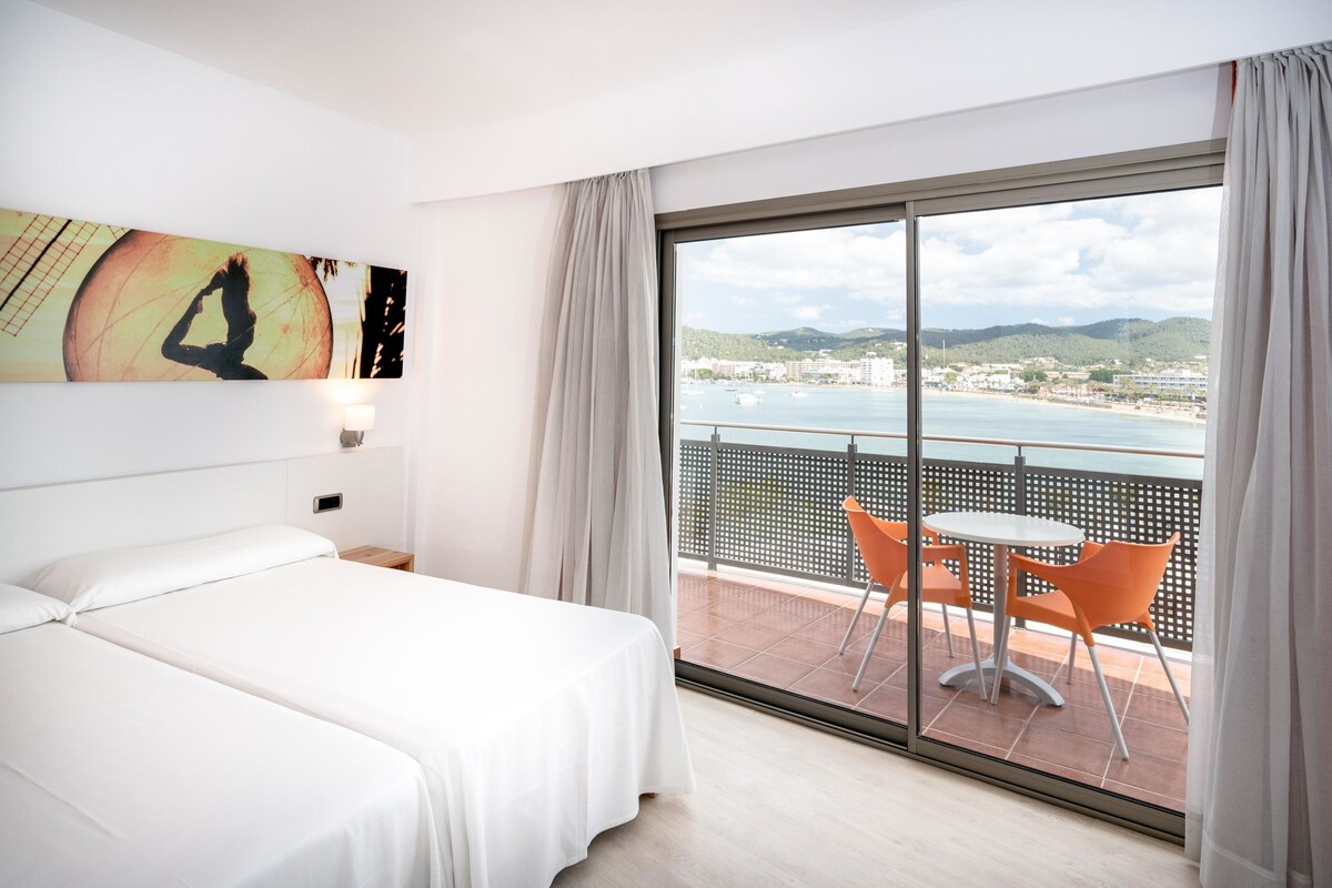 A hotel room with a large bed, a balcony with orange chairs, and a view of the sea and hills.