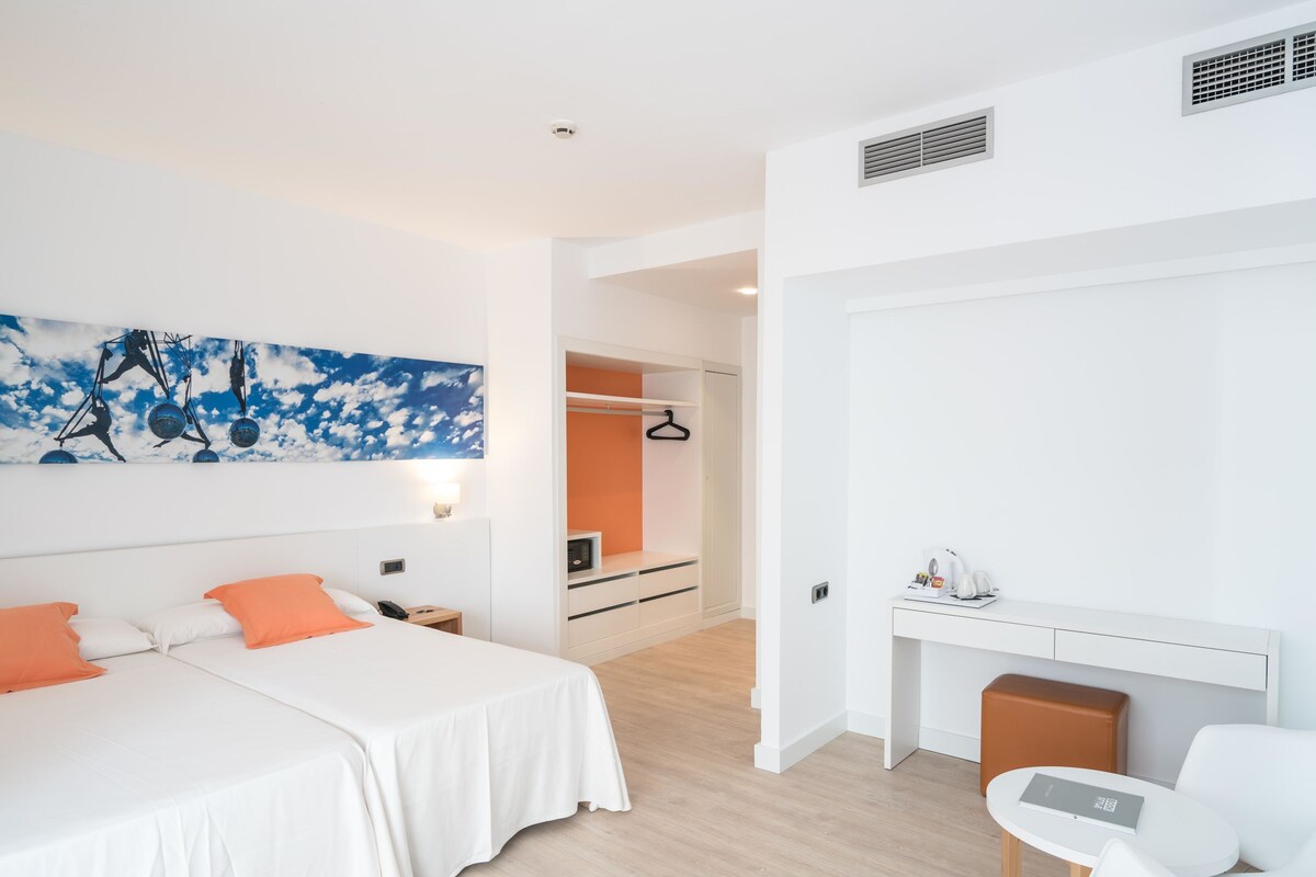 A modern hotel room with two beds, a desk, and a chair. There is a painting on the wall and a closet with an orange interior.
