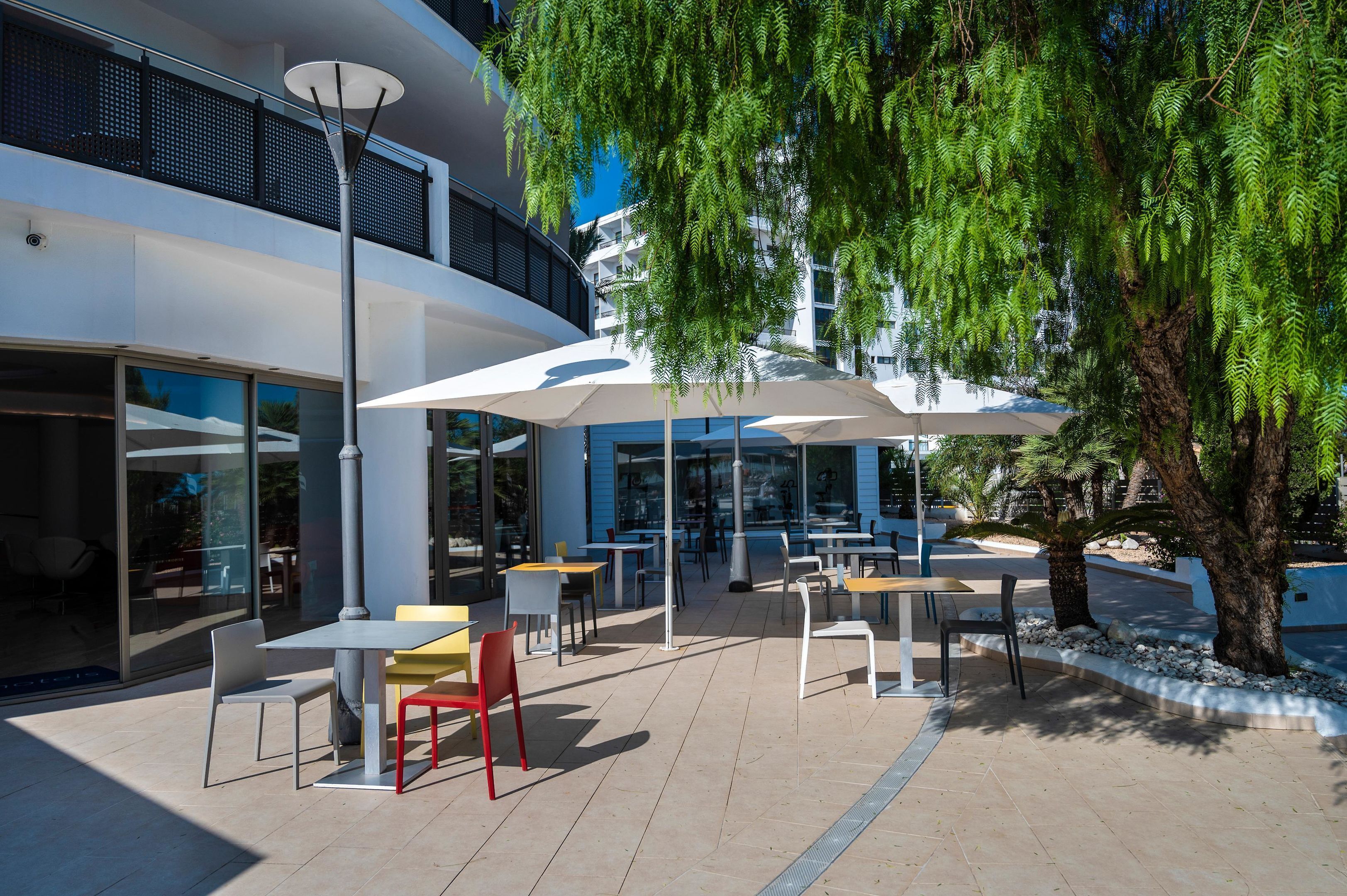 Un espace extérieur aménagé avec des tables et des chaises, entouré d’arbres et bordé d’un bâtiment moderne.