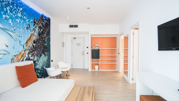 A modern living room with a white sofa, a red cushion, and a wooden coffee table. There is a TV mounted on the wall and a large window with a view of a cityscape. - Hotel THB Ocean Beach - Adults Only (Sant Antoni de Portmany)