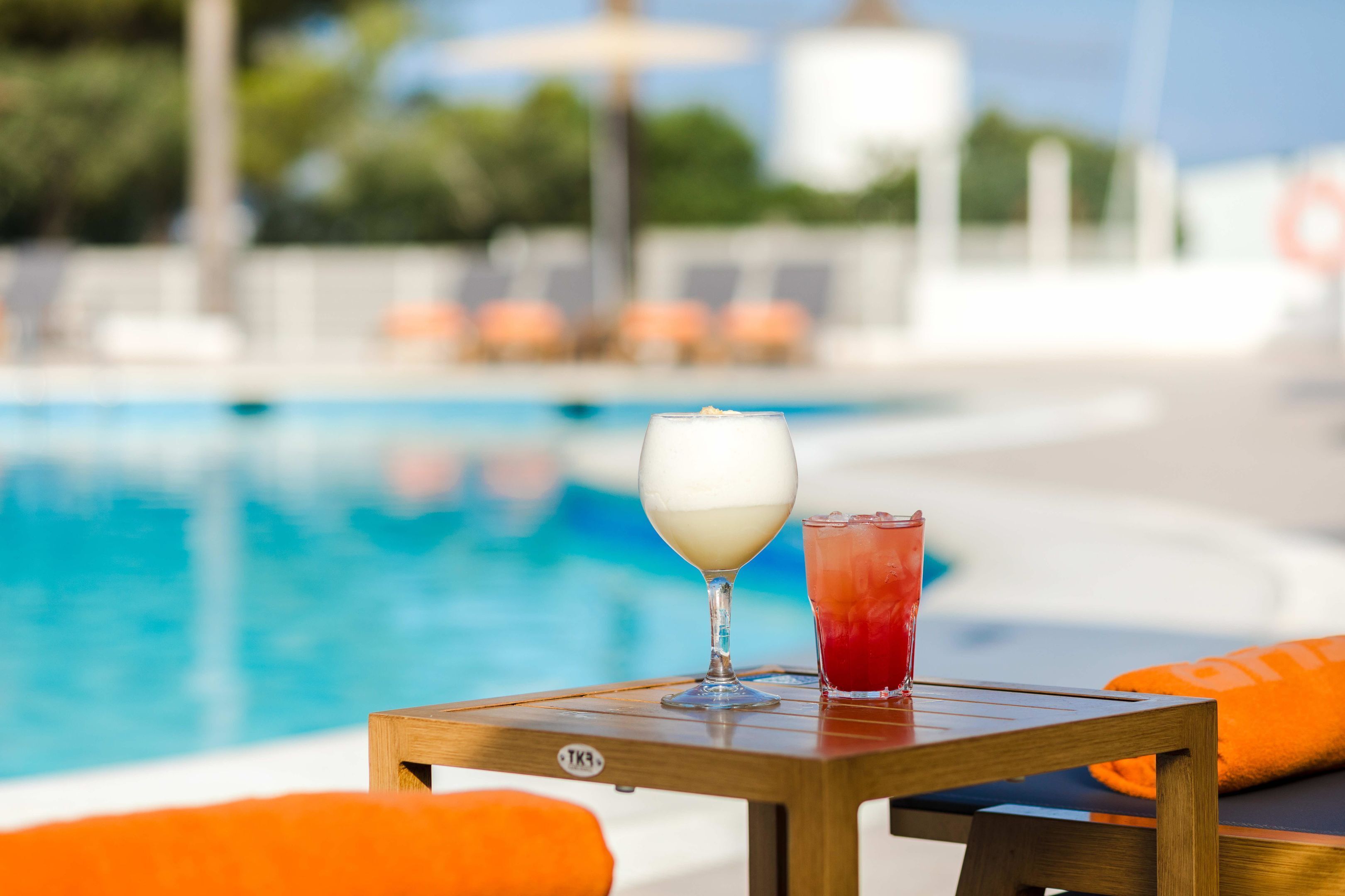 A poolside table with a glass of white drink and a red drink, surrounded by orange cushions.