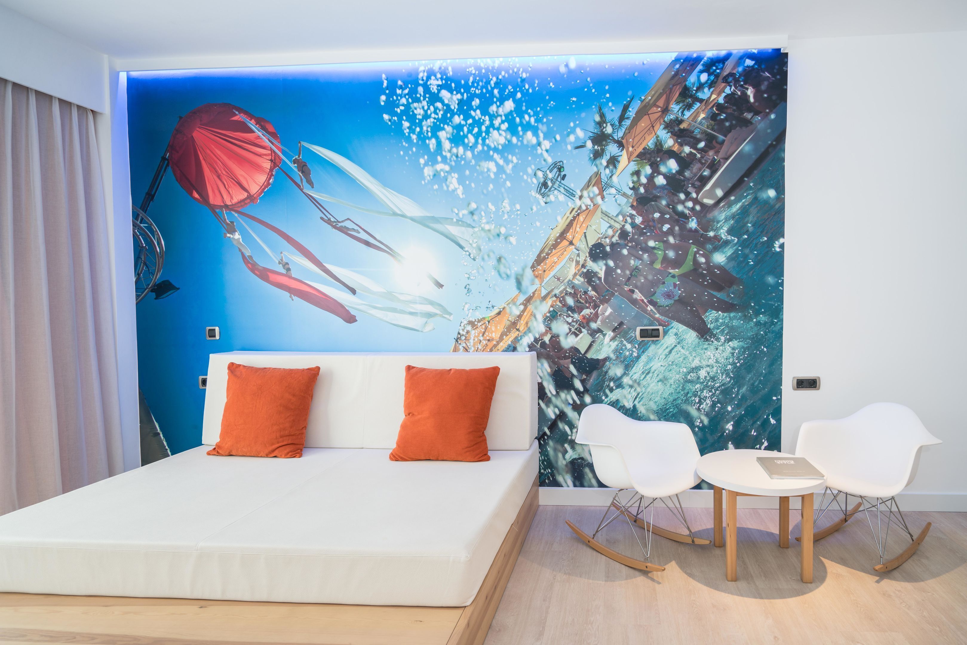 A modern hotel room with a large mural of an underwater scene, a white bed with orange pillows, and two white chairs with a small table.