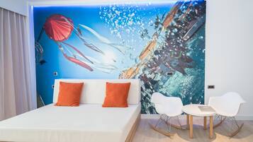 A modern hotel room with a large mural of an underwater scene, a white bed with orange pillows, and two white chairs with a small table.