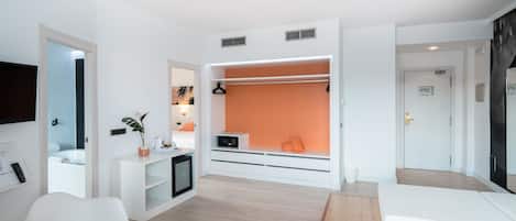 A modern hotel room with a white and orange color scheme, a seating area with a table, a TV mounted on the wall, and a wardrobe with a mirror.