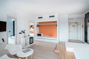 Una habitación de hotel moderna con una decoración en blanco y naranja, un área de descanso con una mesa, un televisor montado en la pared y un armario con espejo.