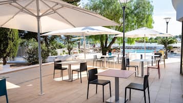 Un patio al aire libre con mesas y sillas bajo sombrillas grandes, con vistas a una piscina y follaje.