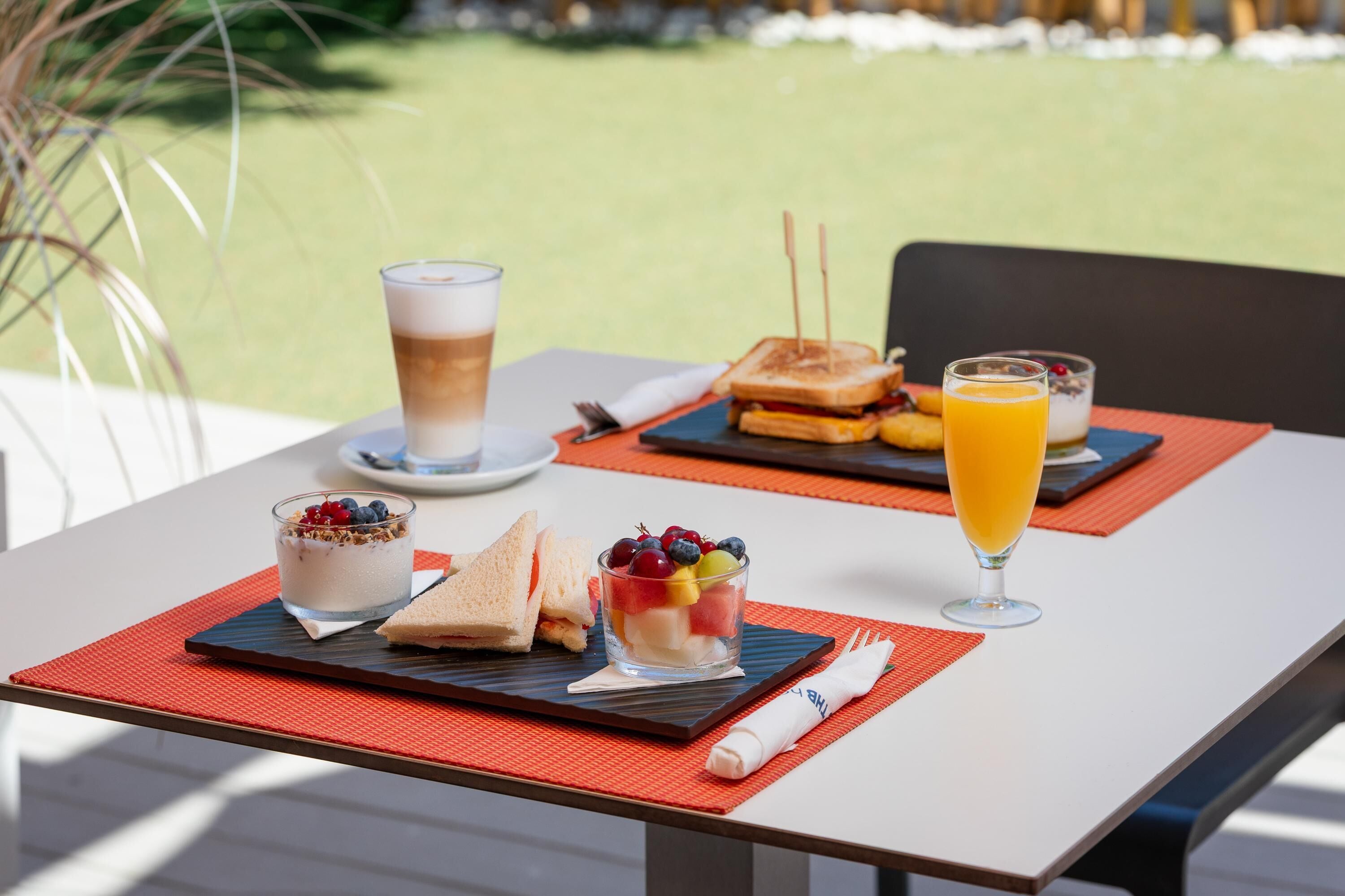 Un desayuno con un sándwich, un bol de fruta y un vaso de zumo de naranja sobre una mesa.