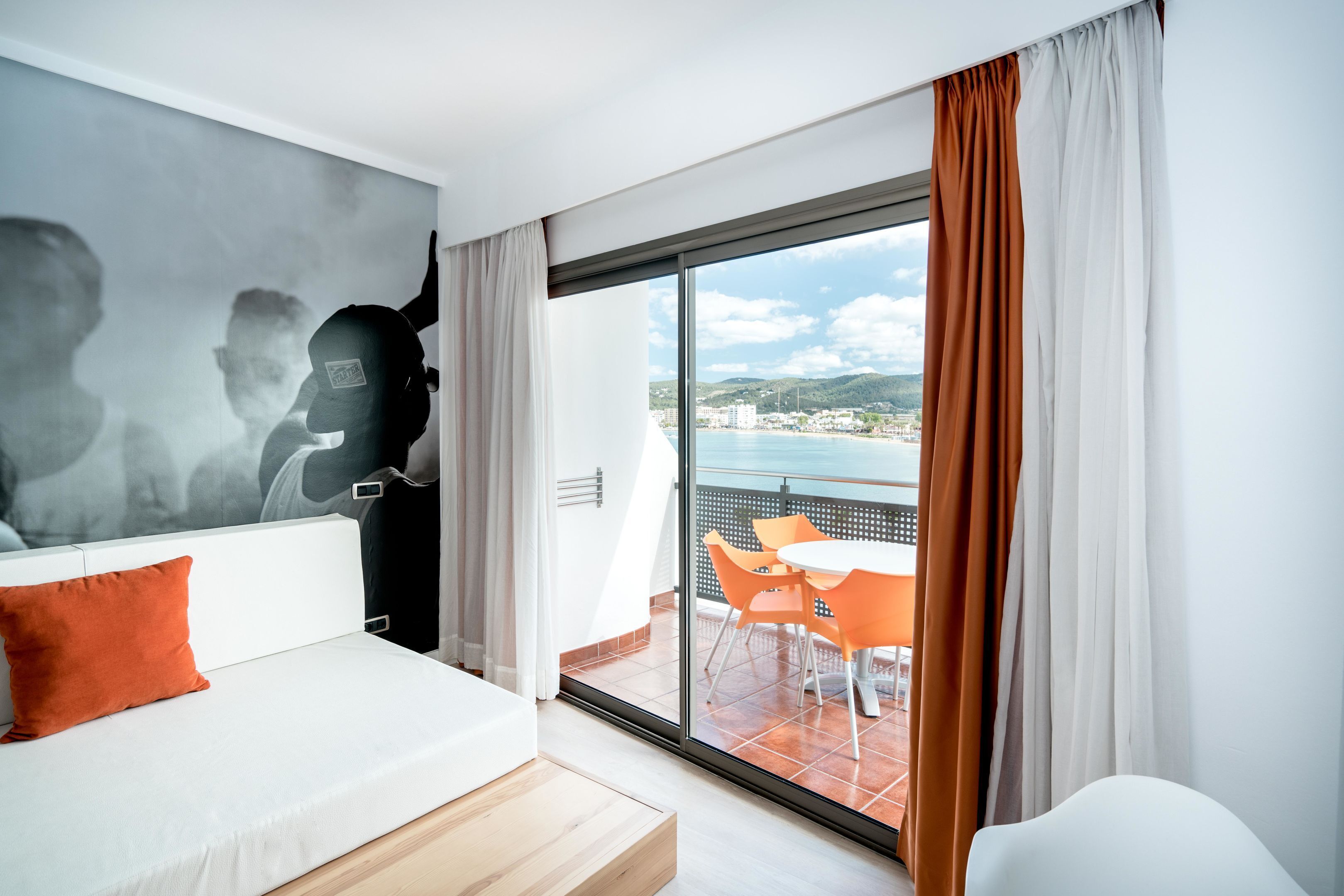 A modern hotel room with a large balcony, a dining area with orange chairs, and a view of the sea and hills.