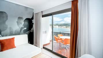 A modern hotel room with a large balcony, a dining area with orange chairs, and a view of the sea and hills.