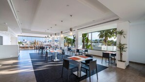 A spacious dining area with modern tables and chairs, large windows offering a city view, and a ceiling with recessed lighting. - Hotel THB Ocean Beach - Adults Only (Sant Antoni de Portmany)