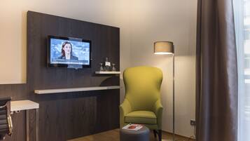 Ein modernes Hotelzimmer mit einem an der Wand befestigten Fernseher, einem grünen Sessel, einem kleinen Hocker mit einer Zeitschrift, einer Stehlampe und einem Holztresen.