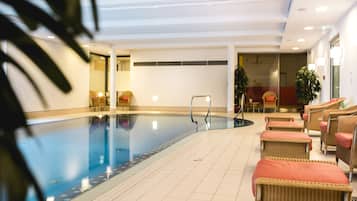 An indoor swimming pool with a tiled floor, wicker chairs with cushions, and potted plants.
