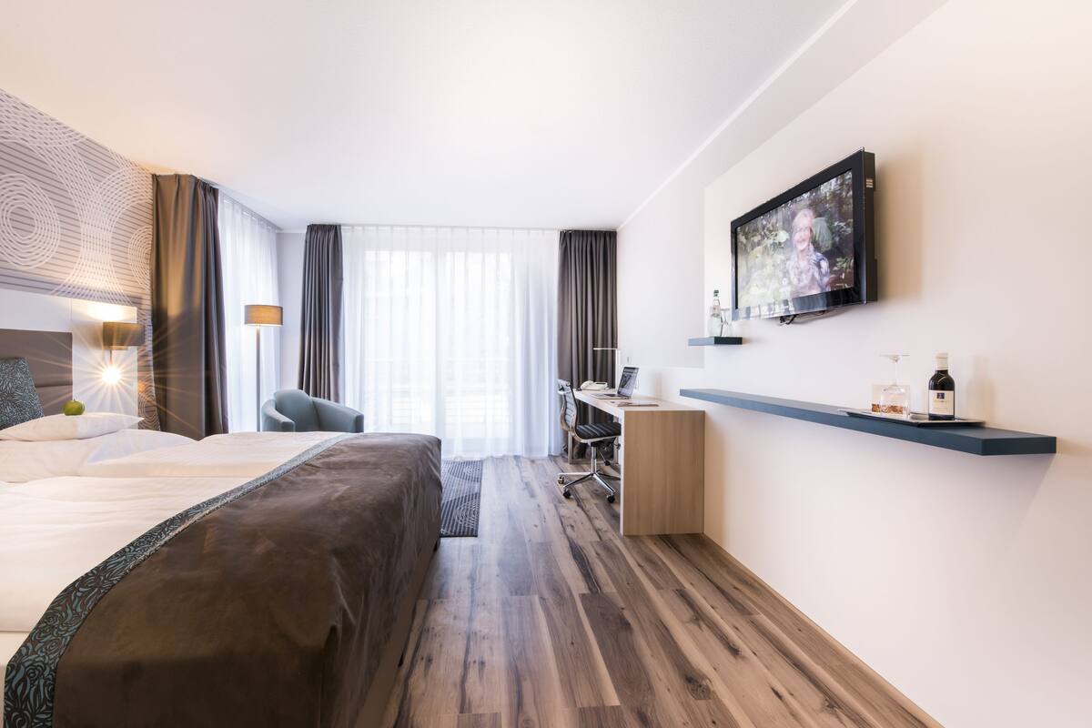 Ein modernes Hotelzimmer mit einem großen Bett, einem Schreibtisch, einem Sessel, einem an der Wand befestigten Fernseher und Holzboden.