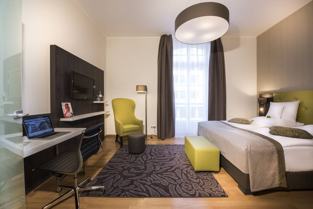 Ein modernes Hotelzimmer mit Schreibtisch, Laptop, grünem Sessel und einem Bett mit grünem Kopfteil.