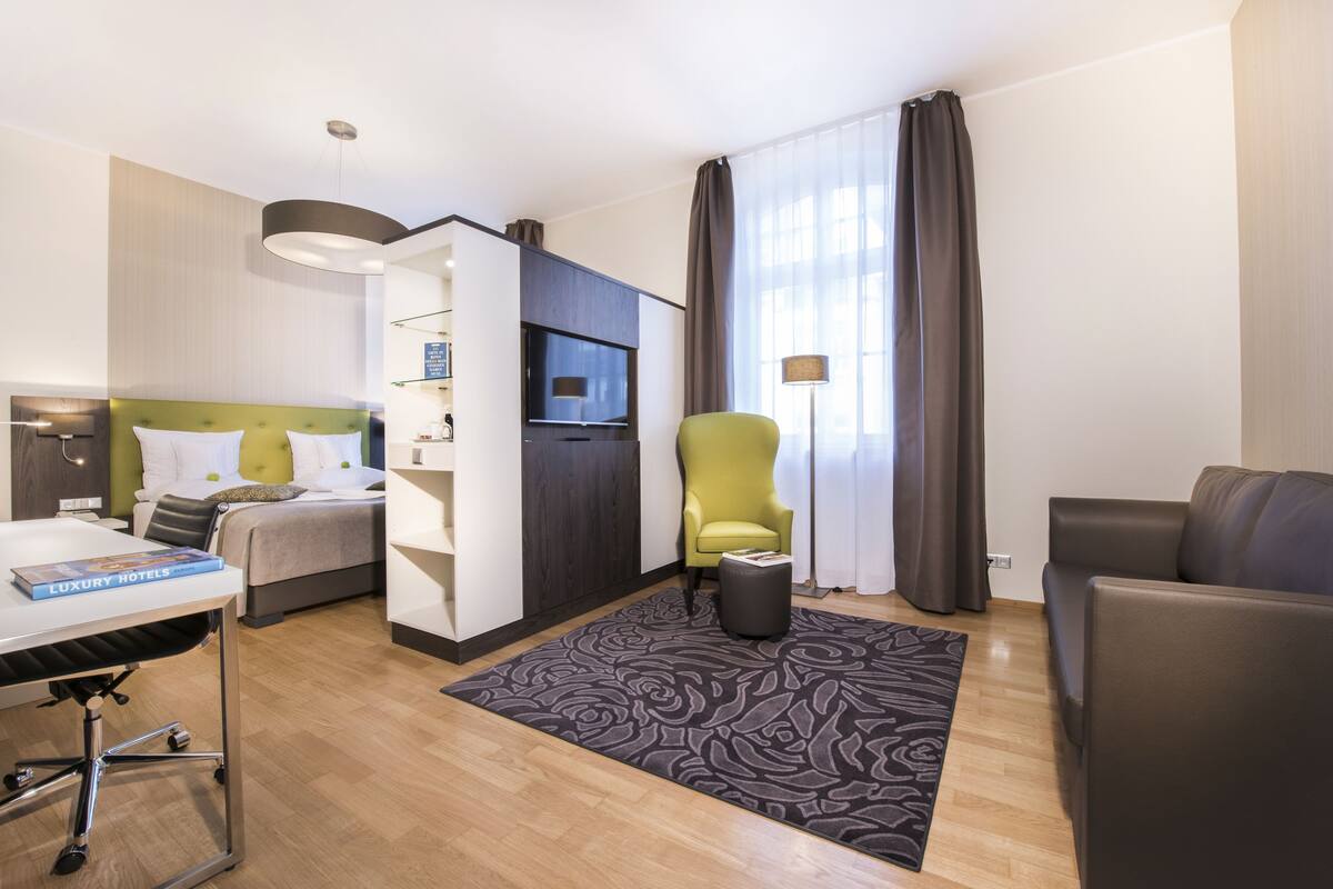 Ein modernes Hotelzimmer mit Bett, Schreibtisch, Sessel, Sofa, Fernseher und einem großen Fenster mit Vorhängen.