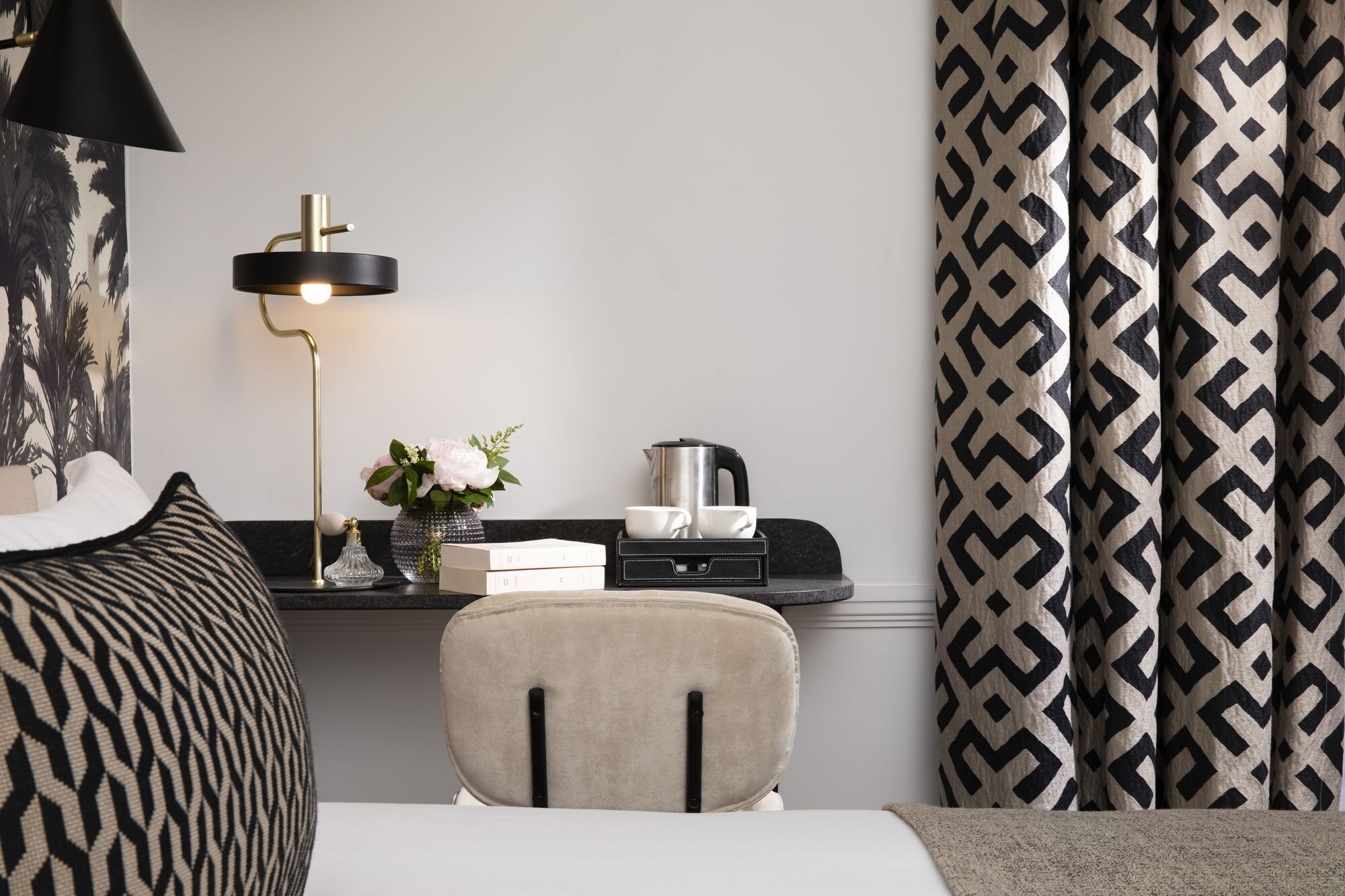 A hotel room with a bed, a desk with a lamp, a chair, a coffee maker, and patterned curtains.