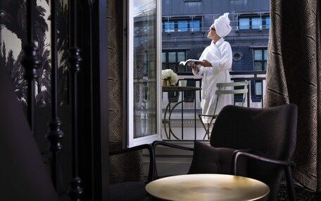 A room with a large window, a chair, a table, and a person reading a book.