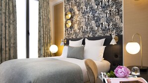 Una cama bien hecha con una manta gris, un cabecero con estampado floral, dos lámparas de noche y una mesita de noche con un libro y una flor.