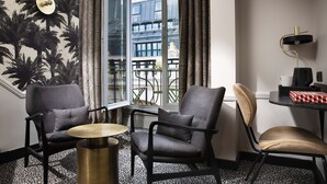 Una habitación de hotel moderna con un ventanal grande, dos sillones, una mesita redonda, un escritorio negro con una lámpara y una alfombra estampada.