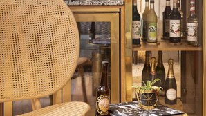 Una zona de bar con una silla de mimbre, una mesa redonda dorada y una vitrina de cristal llena de botellas de licores.