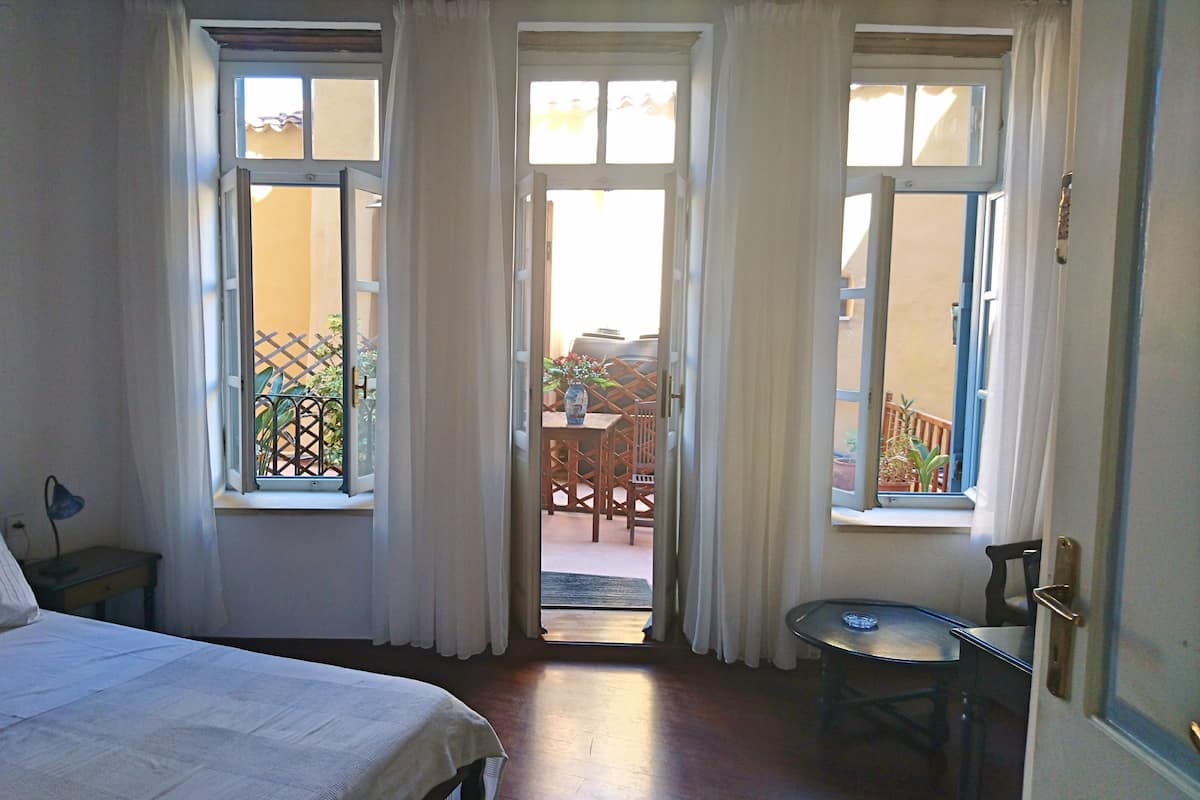 A bedroom with a bed, a small table, and two open windows leading to a balcony.
