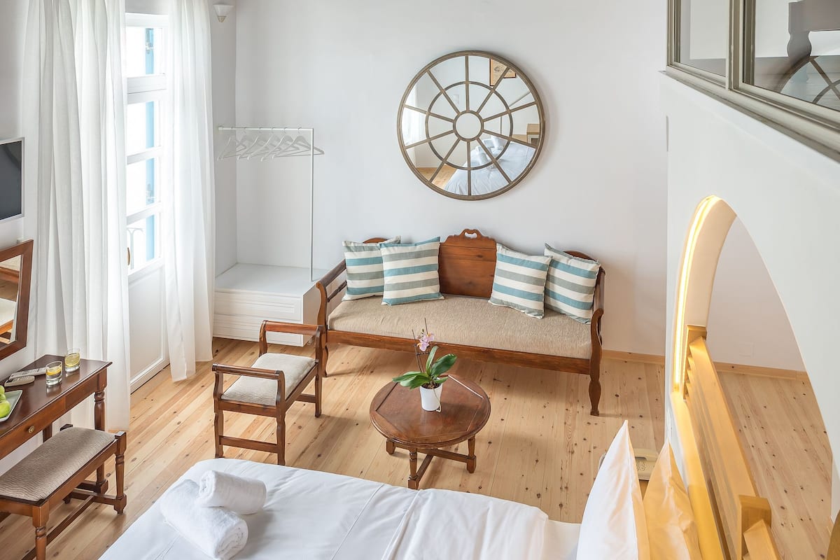 A hotel room with a wooden ceiling, a bed with white linens, a wooden bench, a small table with a plant, and a wall-mounted mirror.
