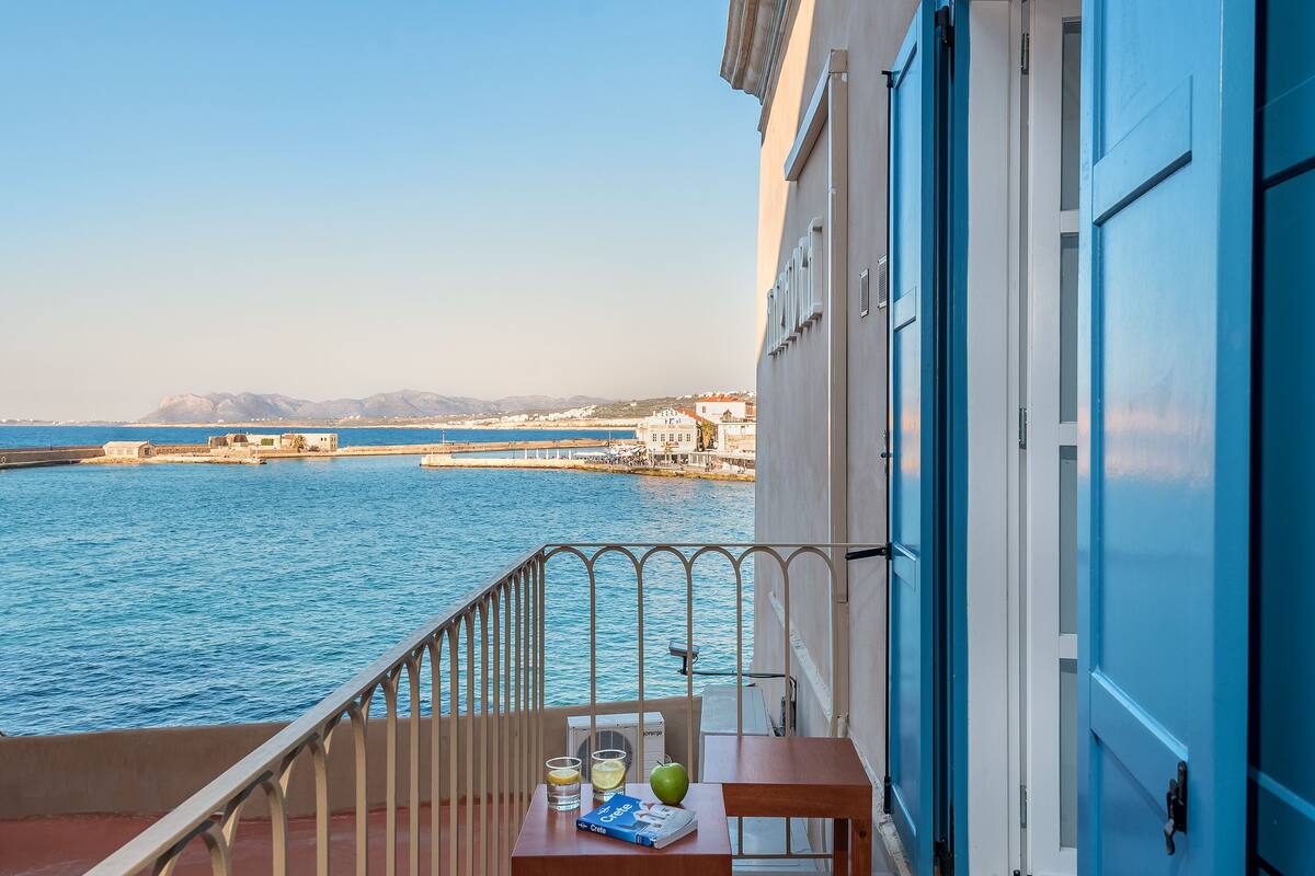 A balcony with a view of the sea and a cityscape. There is a table with a book, glasses, and a fruit.