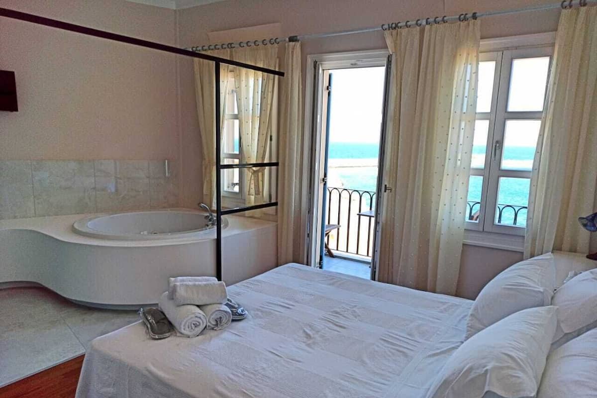 A bedroom with a large bed, a bathtub, and a balcony with a view of the sea.