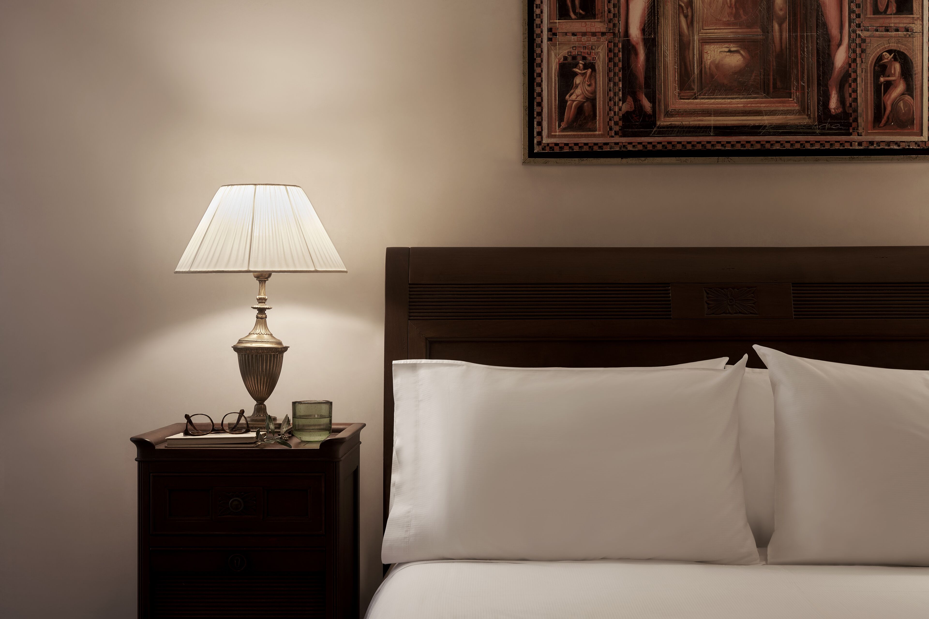 A neatly made bed with white linens, a bedside table with a lamp, glasses, and a glass of water.