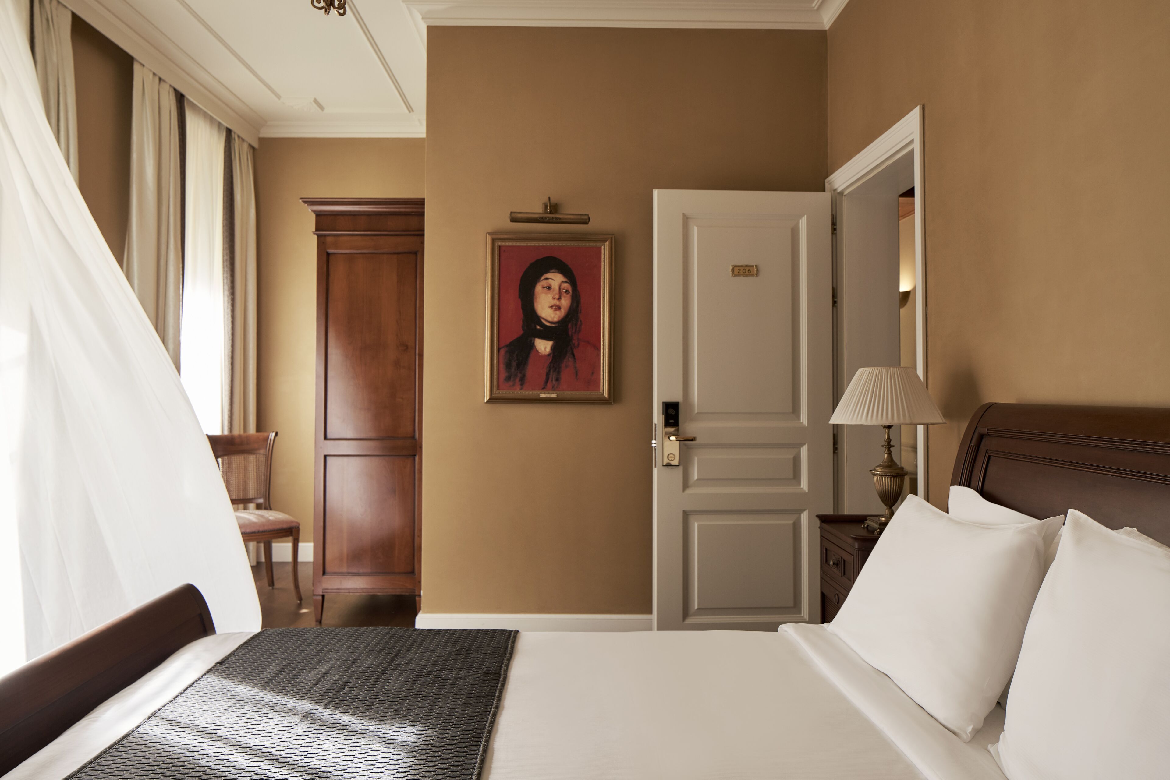 A hotel room with a bed, a wooden wardrobe, a painting on the wall, a door, and a nightstand with a lamp.