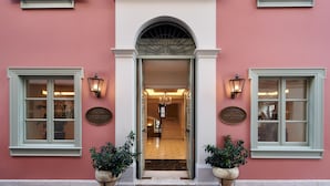 Una entrada de hotel con fachada rosa, un arco central y dos plantas en macetas a la entrada.