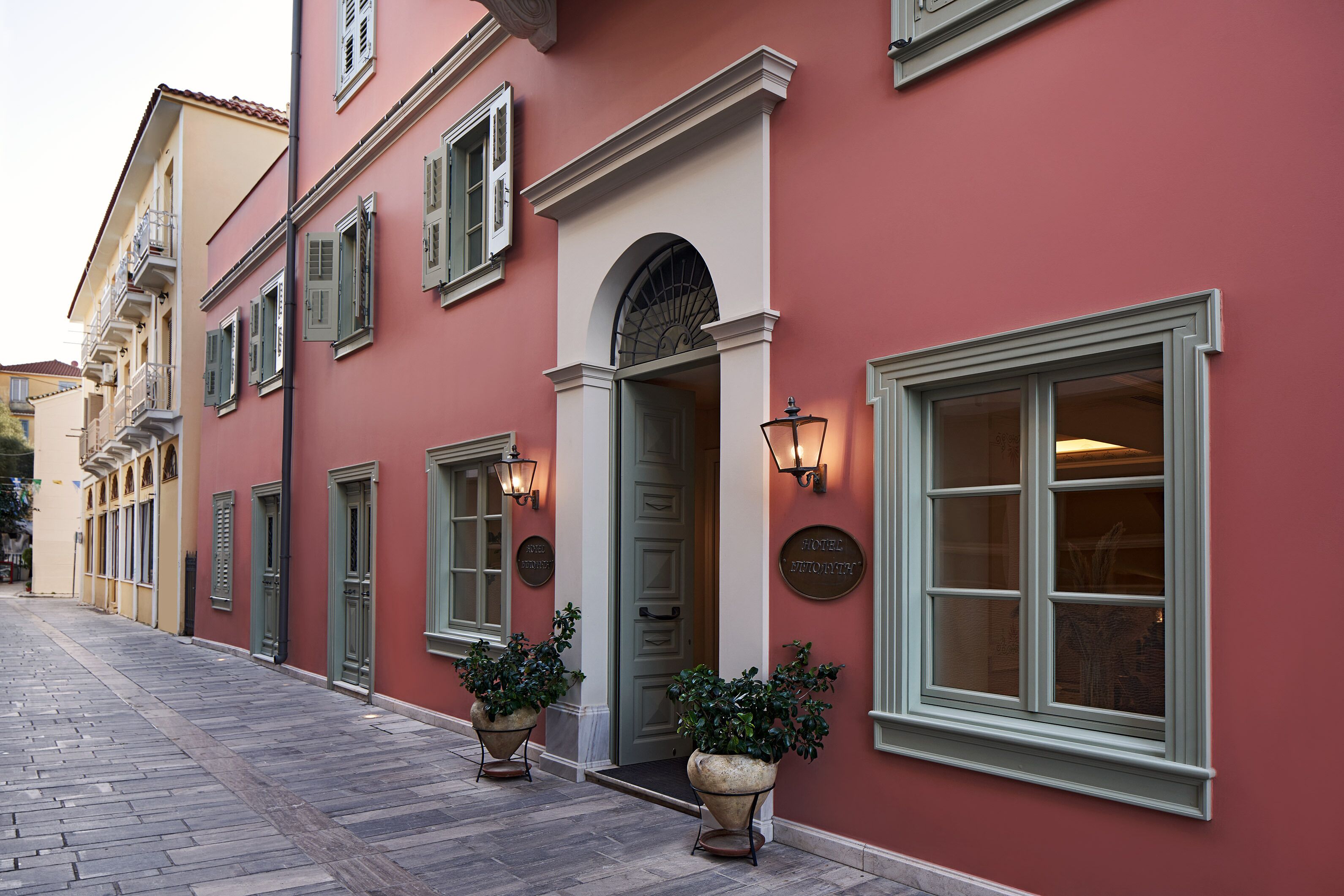 A hotel facade with a red exterior, green shutters, and a prominent entrance with a door and arched window.
