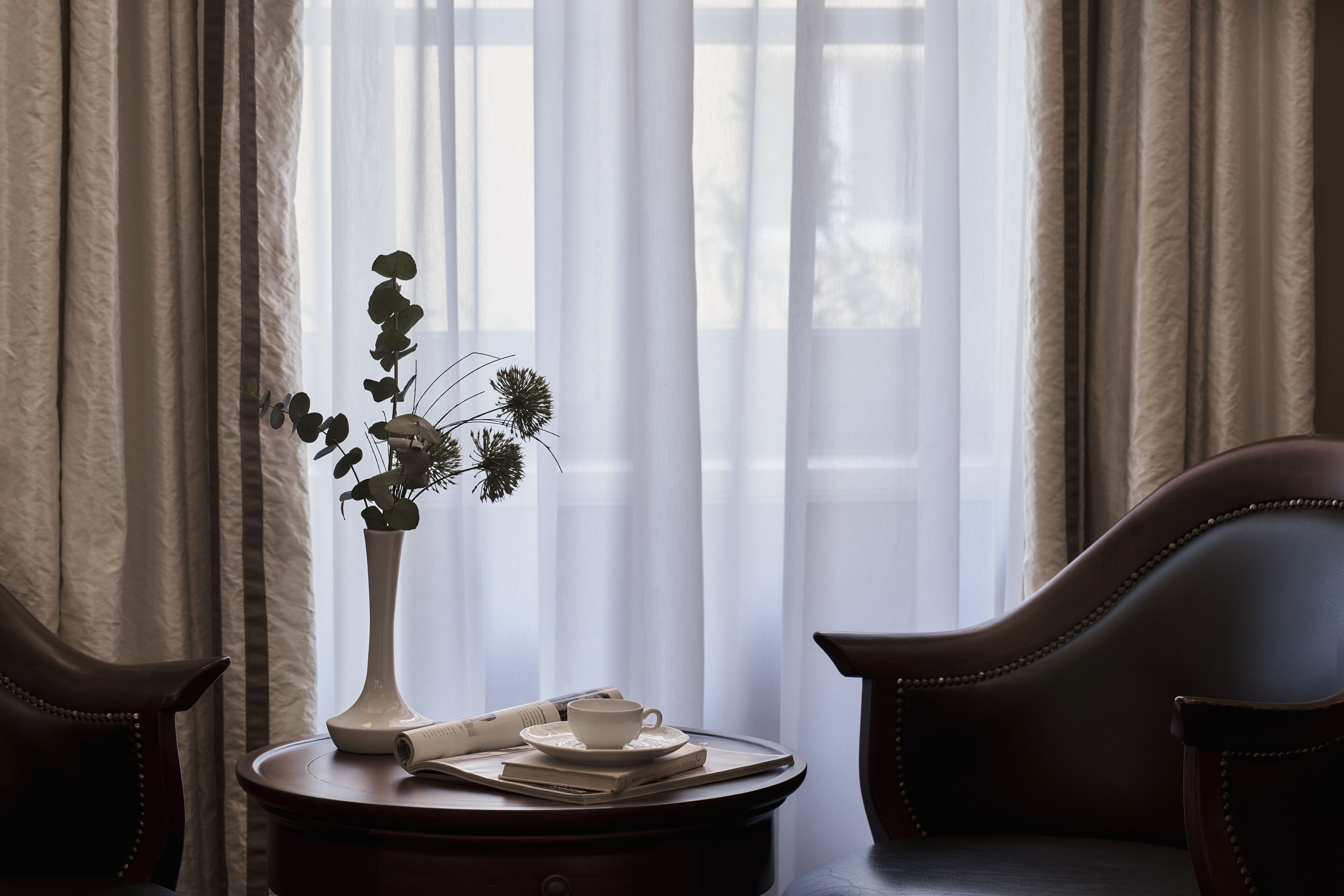 A room with a window, curtains, a vase with flowers, a cup, and a newspaper on a small table.