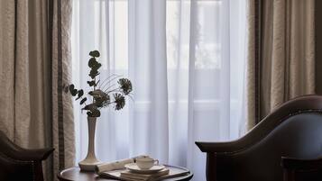 A room with a window, curtains, a vase with flowers, a cup, and a newspaper on a small table.