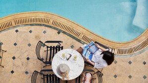 Zona de piscina con mesa y sillas, una persona leyendo un libro y suelo de baldosas.