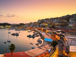 Un pueblo costero al atardecer con un puerto deportivo lleno de barcos, una carretera curva y edificios con letreros iluminados.