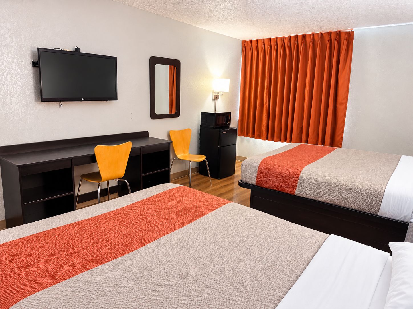 Une chambre d’hôtel avec deux lits, un bureau avec des chaises, une télévision, un miroir et une fenêtre avec des rideaux.