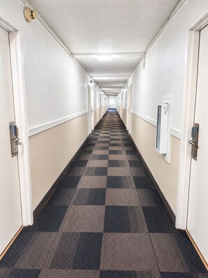 A long, narrow hotel corridor with checkered carpet, white walls, and doors on both sides. - Motel 6 New Stanton, PA (New Stanton)