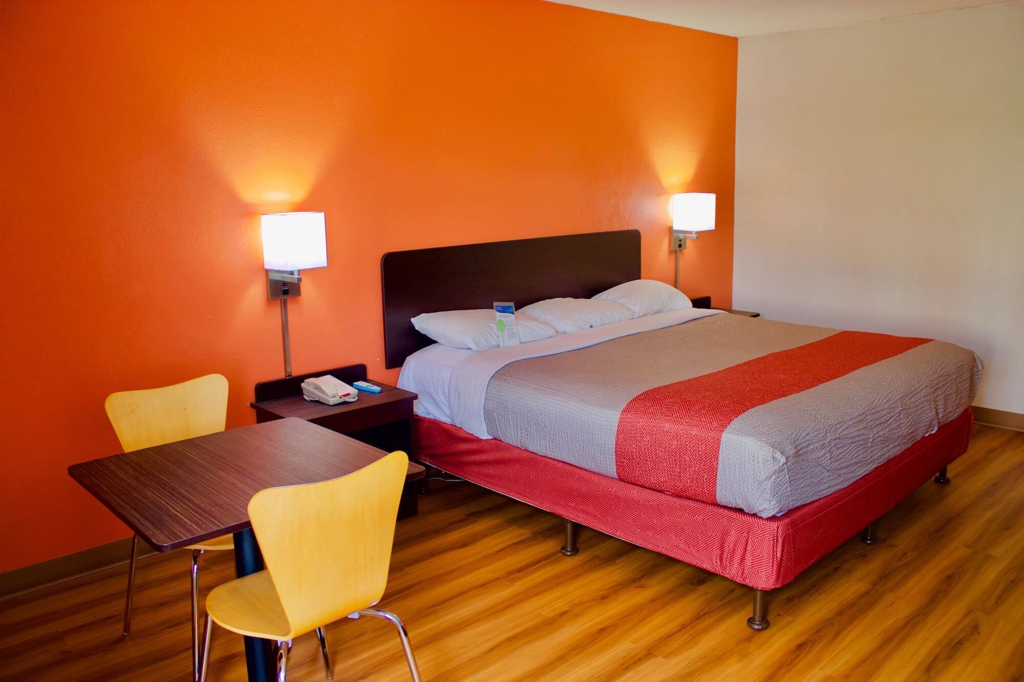 A hotel room with a bed, a wooden nightstand, a chair, and two wall-mounted lamps.