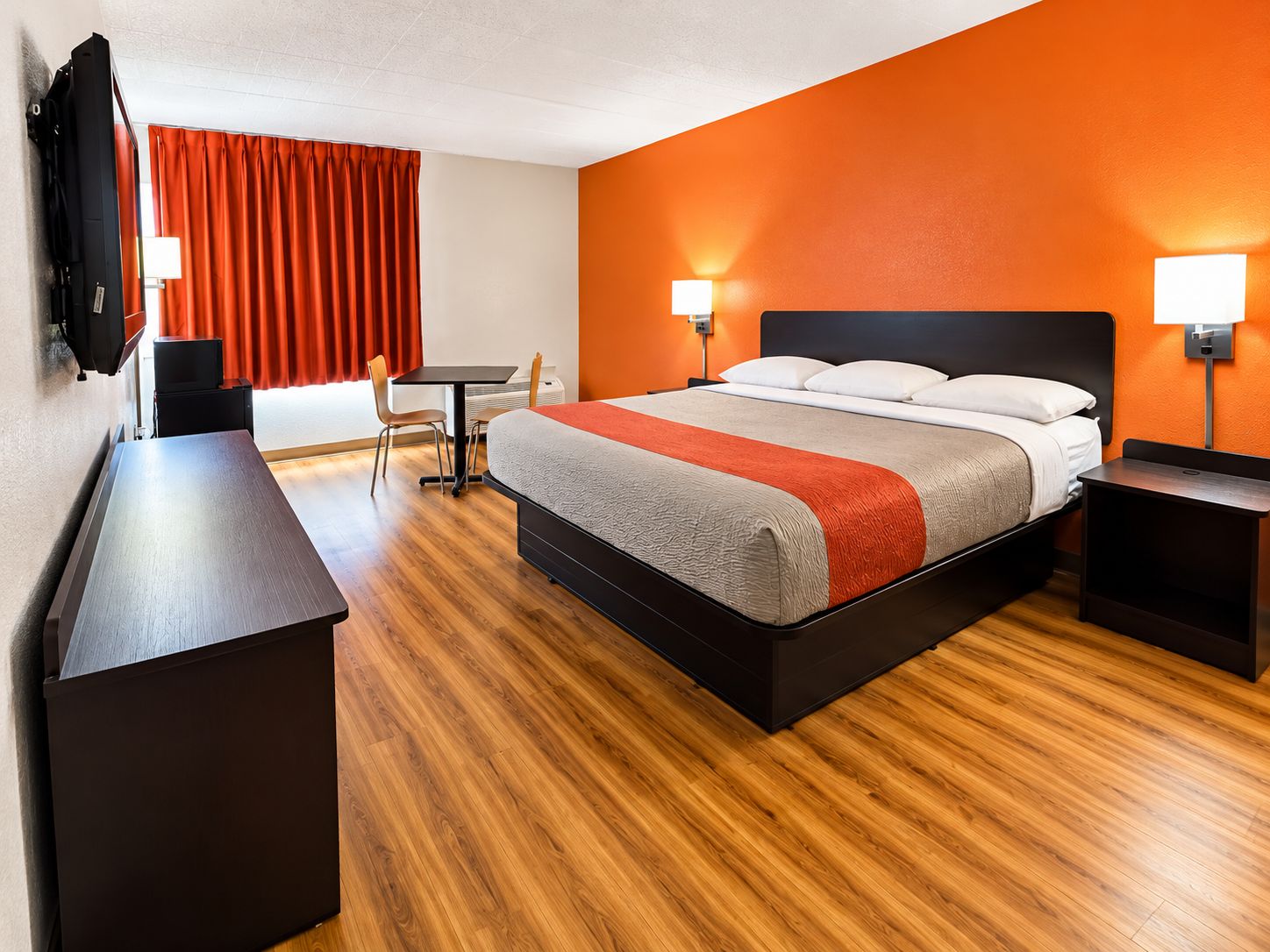 Habitación de hotel con cama, mesitas de noche, escritorio, silla, televisión y cortinas anaranjadas.