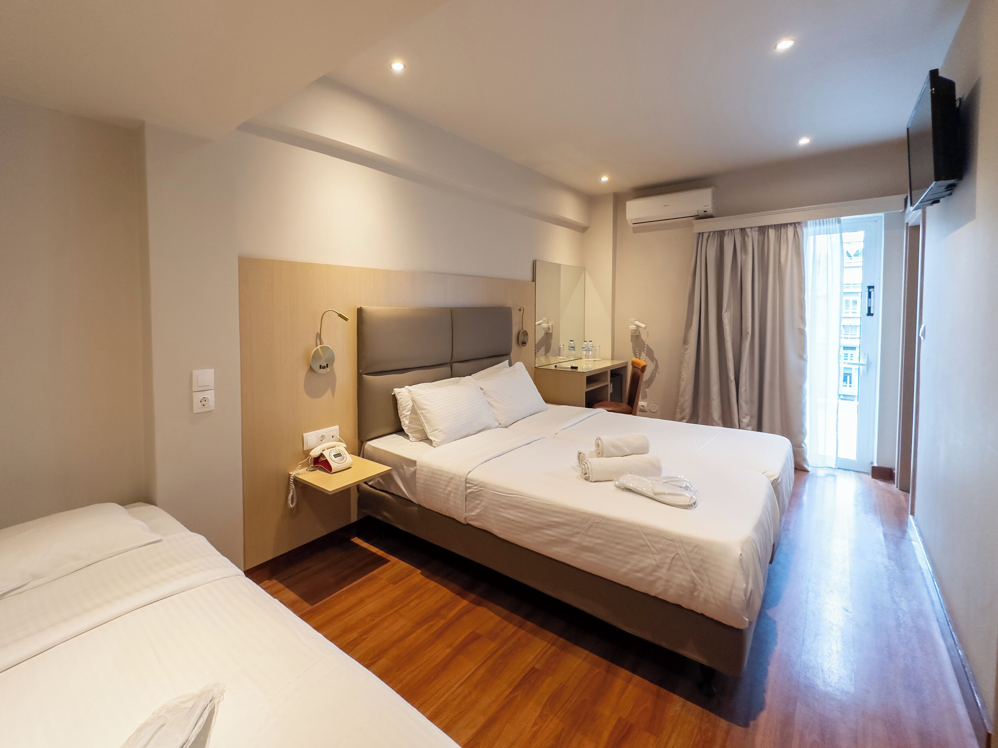 A hotel room with a large bed, a smaller bed, a bedside table with a phone, a mirror, and an air conditioning unit.