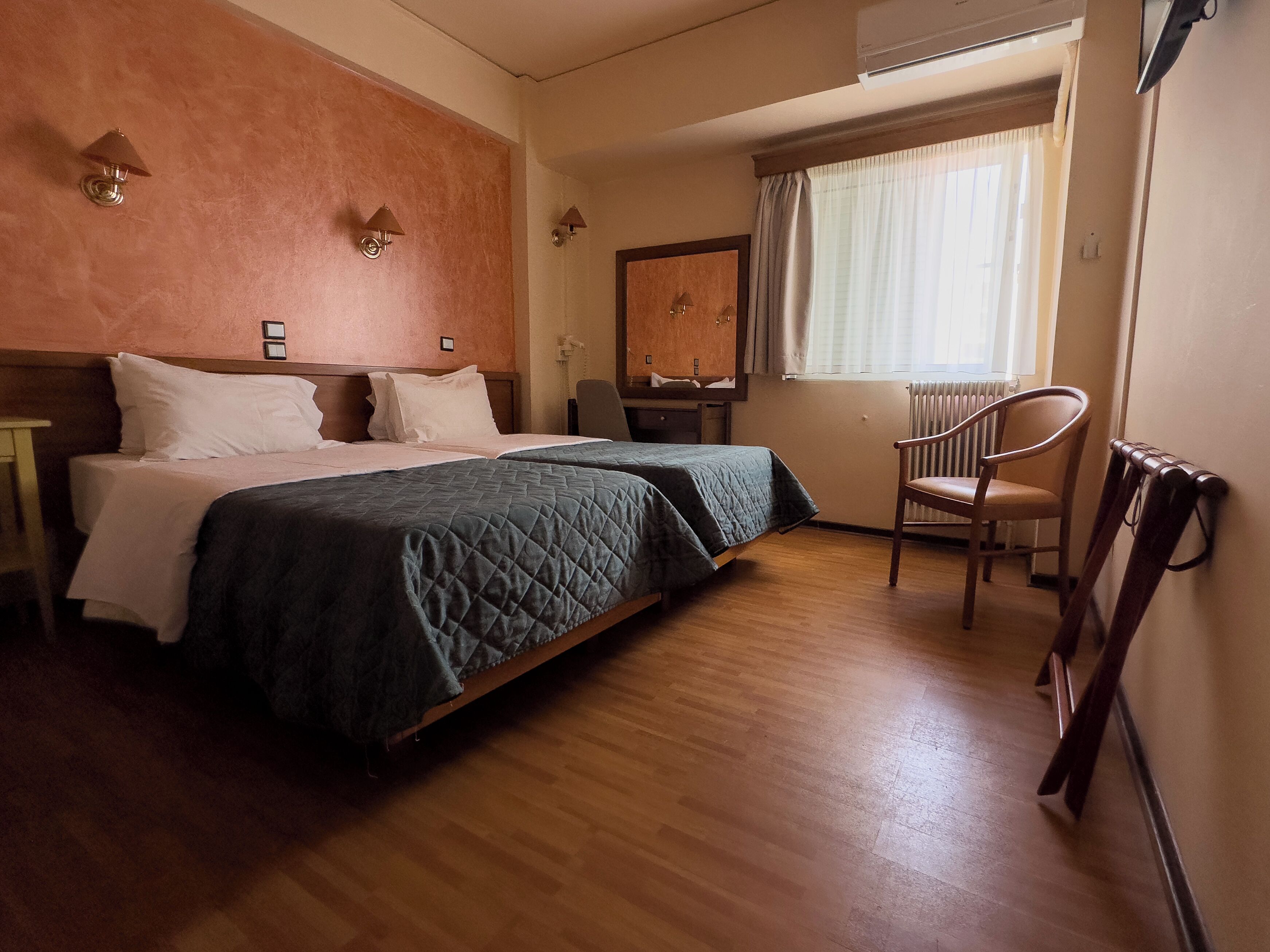 A hotel room with two beds, a wooden floor, a chair, a desk, and a window with curtains.