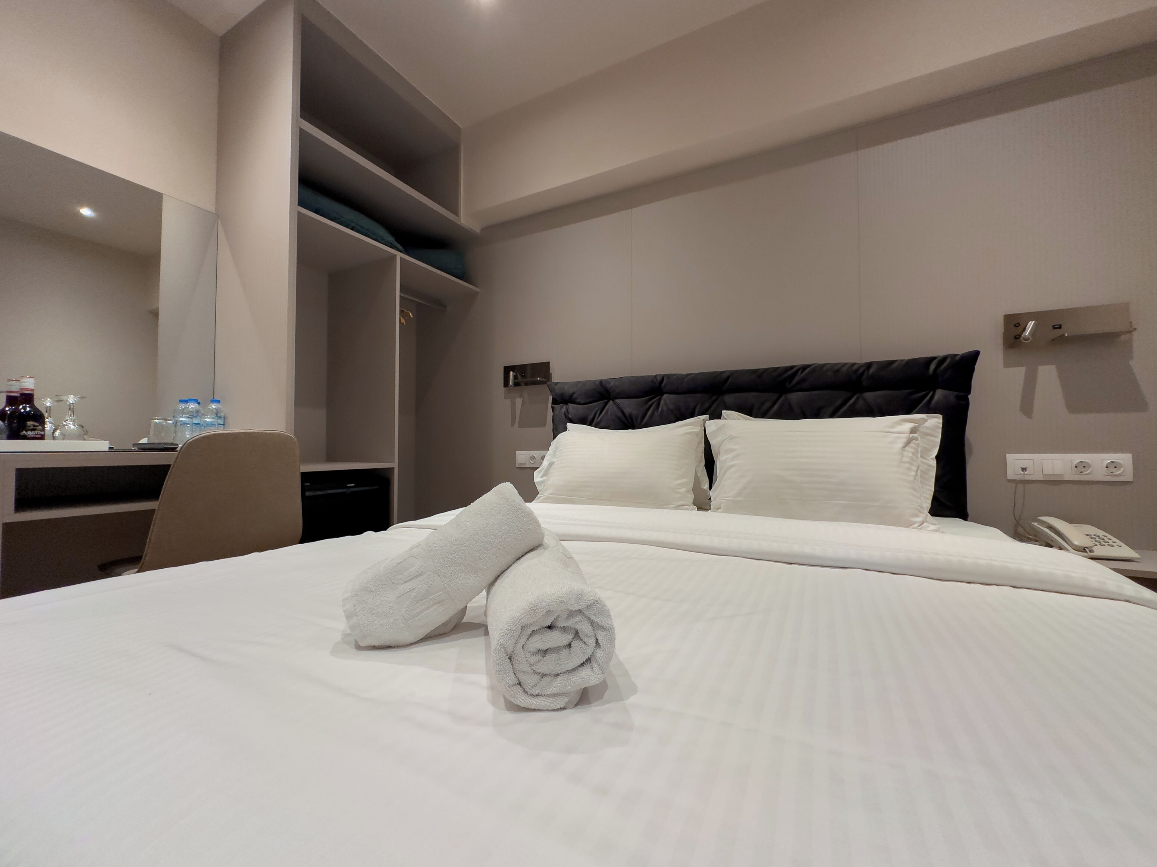 A hotel room with a bed, a desk, a chair, a mirror, and a wall-mounted phone.