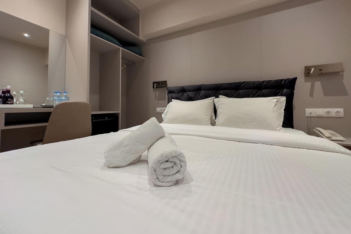 A hotel room with a bed, a desk, a chair, a mirror, and a wall-mounted phone.