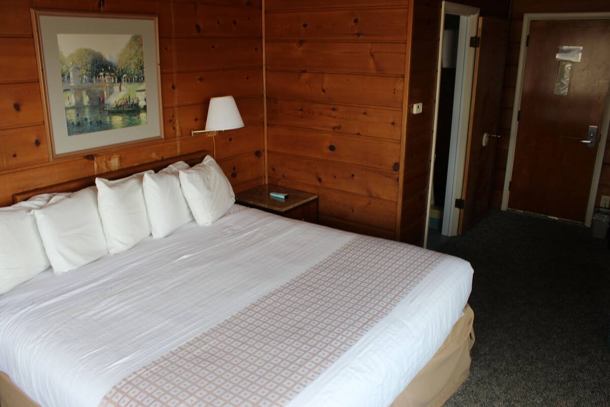 A hotel room with a large bed, a nightstand with a lamp, and a door leading to another room.