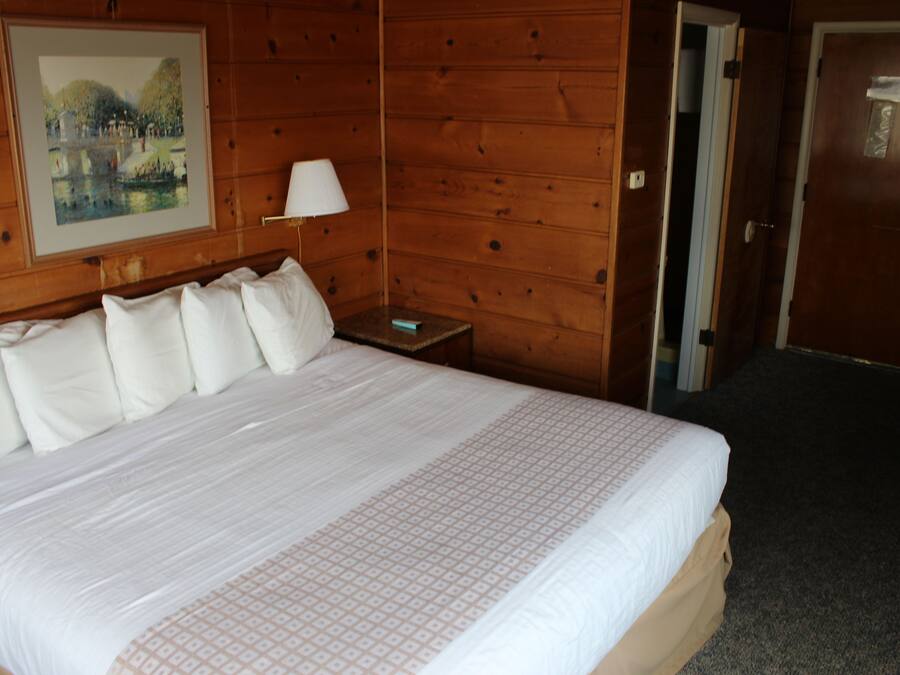 Habitación de hotel con una cama grande, una mesita de noche con lámpara y una puerta que da a otra habitación.
