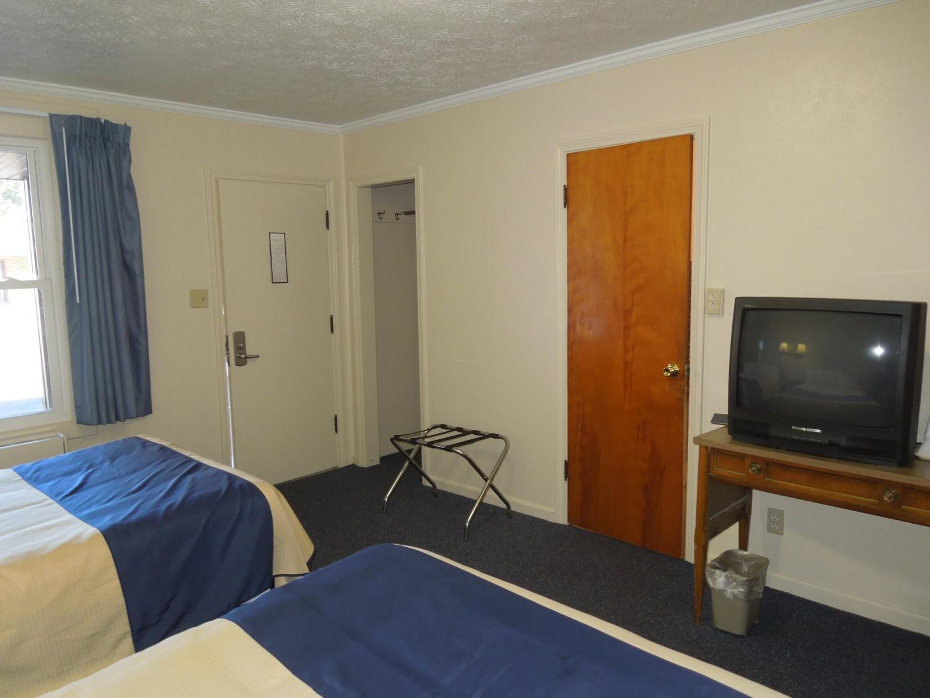 A hotel room with two beds, a television on a wooden table, a door, and a window with curtains.