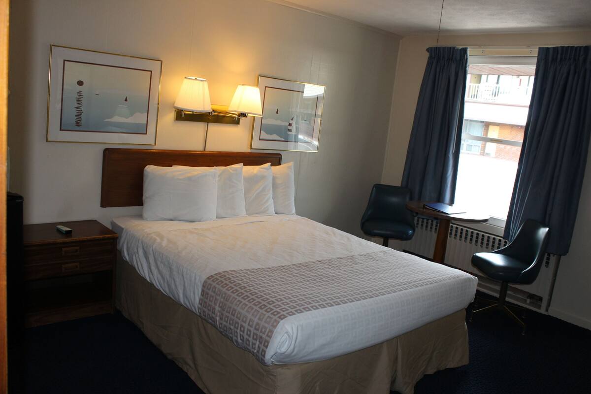 A hotel room with a bed, bedside table, lamp, chair, and two framed pictures on the wall.