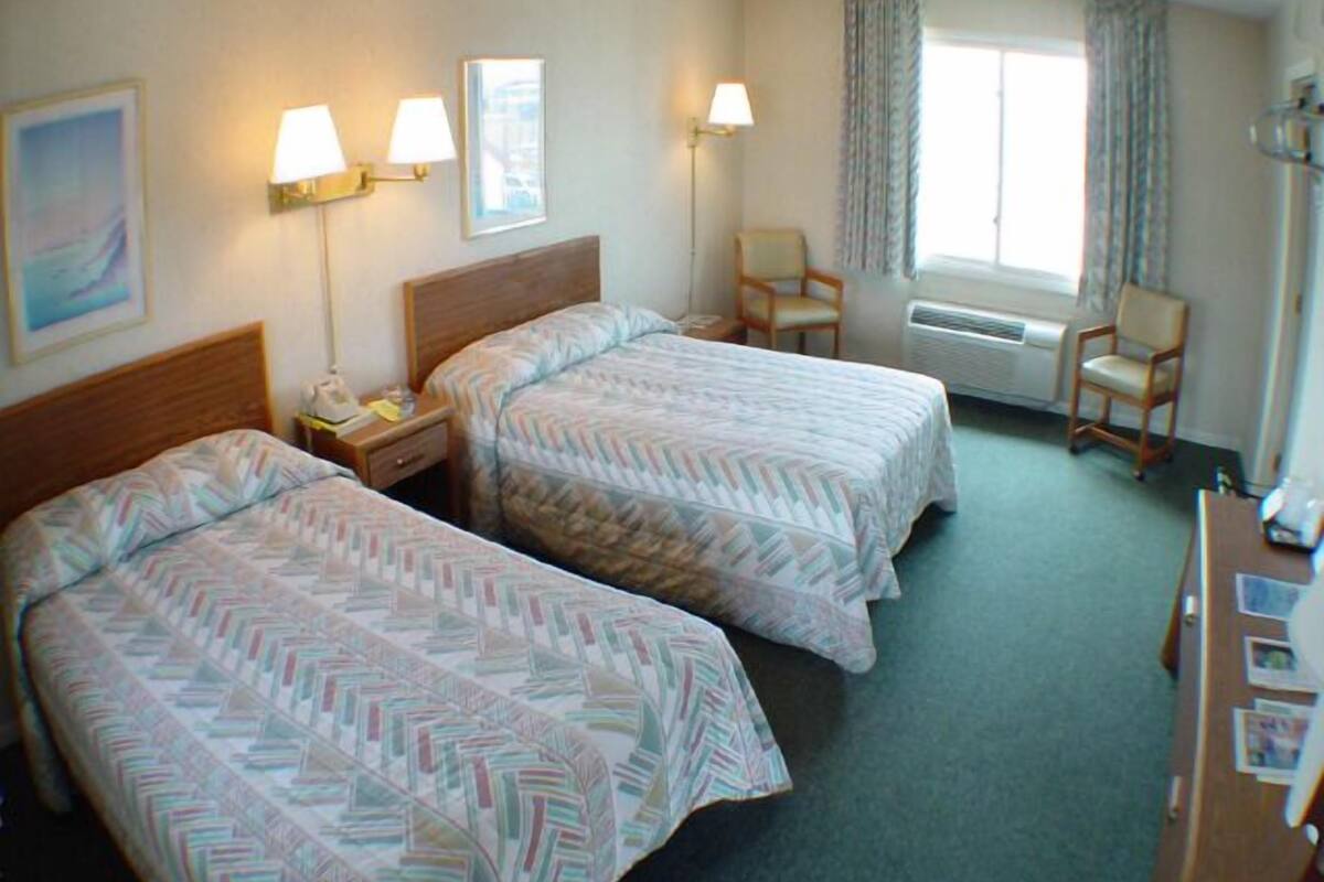 A hotel room with two beds, a desk, a chair, a window with curtains, and wall-mounted lamps.