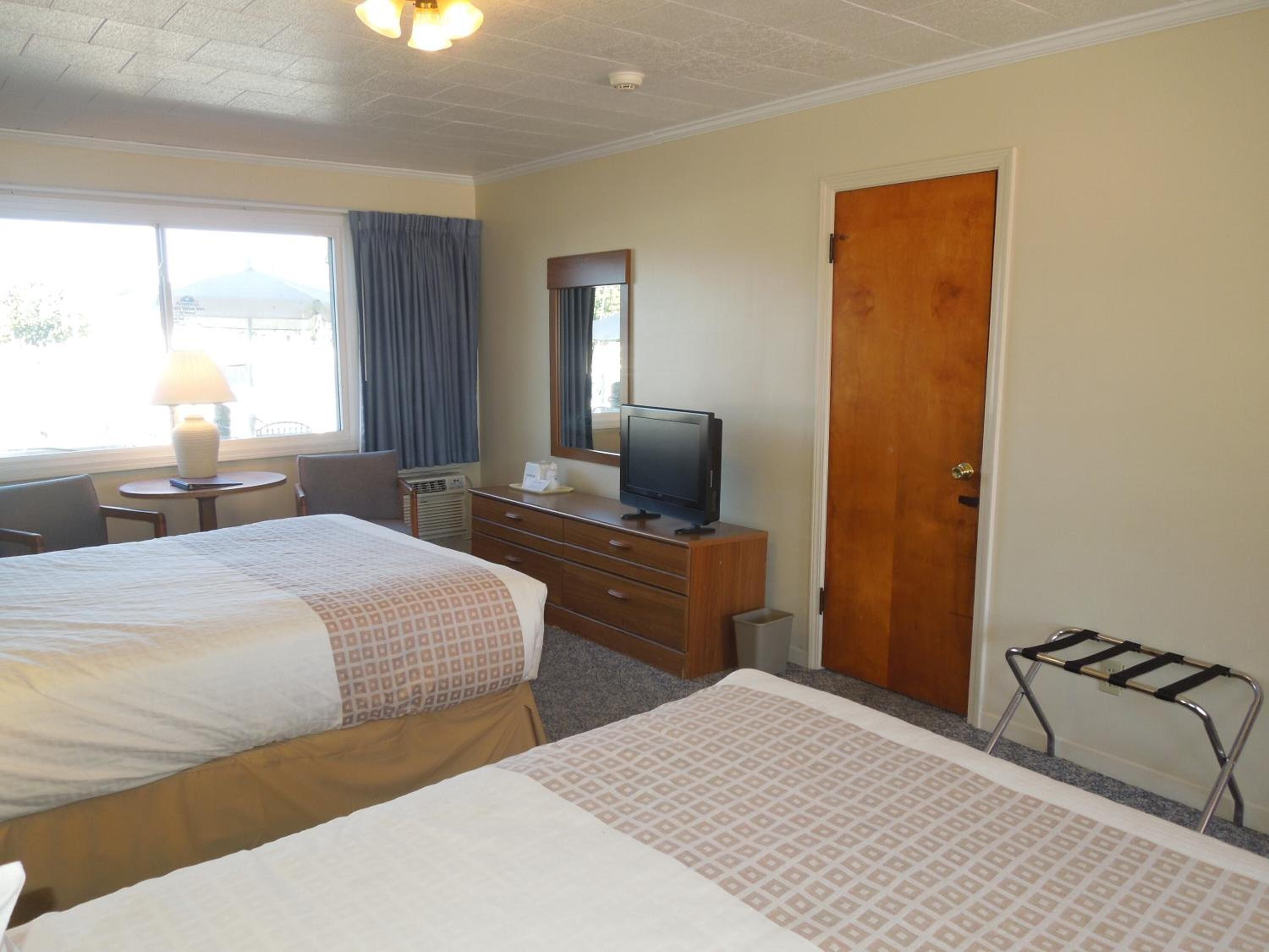A hotel room with two beds, a desk with a lamp, a television on a dresser, and a door.